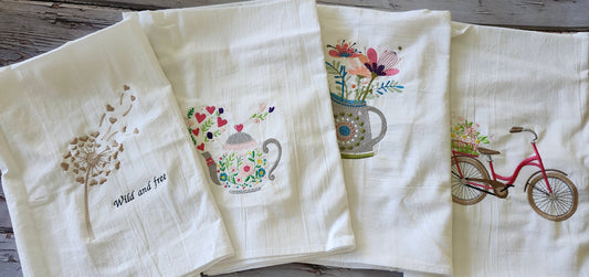Four white flour sack tea towels side-by-side featuring colorful embroidery of a teapot, bicycle, dandelion, and flowers on a rustic wooden background
