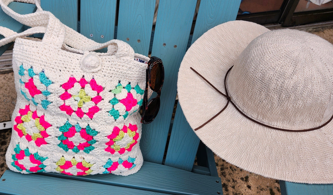 A white handmade crochet tote bag with colorful neon pink, turquoise, and yellow granny squares resting on a blue wooden chair next to a sun hat and sunglasses.