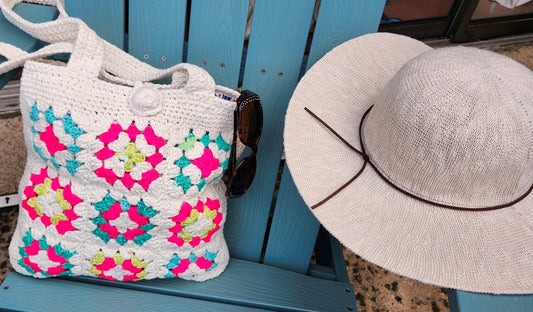 A white handmade crochet tote bag with colorful neon pink, turquoise, and yellow granny squares resting on a blue wooden chair next to a sun hat and sunglasses.