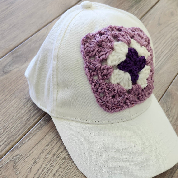 White cap with a purple and white crocheted square on a wooden surface